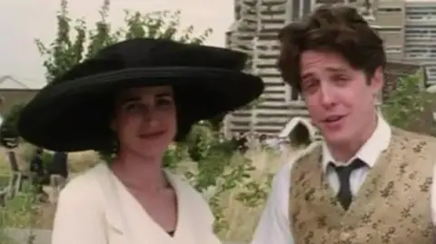 Four Weddings and a Funeral/Polygram Film Production/Working Title/Mike Newell A woman (Andie MacDowell) wearing a large black hat and a white suit jacket standing next to a man (Hugh Grant) wearing a white shirt, black tie and golden embellished suit vest. They are standing outside grasslands and a tall building to the right.