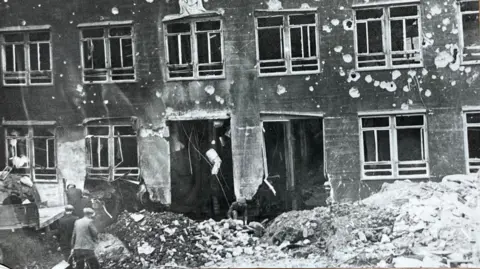 Coventry Archives A black and white photograph of a building damaged by bombing. There is debris on the ground and holes in the walls.
