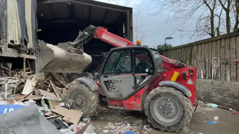 A red telehandler vehicle is central, positioned in front of metal sheets and wooden refuse in a scrapyard environment 