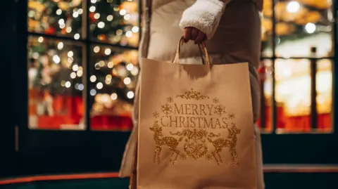 Getty Images A woman wearing a brown coat with white fingerless gloves carries a bag decorated with "Merry Christmas" past a shop lit up for the festive season.