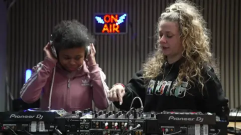 A young girl with dark curly hair in a pink hoodie holds silver headphones on her head as she listens to sounds coming from a mixing board. On the right of the image, an older woman with long blonde curly hair teaches her. A neon sign saying 'on air' can be seen in the background