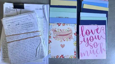 A bundle of handwritten letters tied with an elastic band alongside a selection of cards including anniversary cards and others that say "miss you" and "I love you"