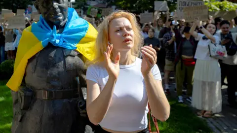 A woman with blonde hair and a whiteT -shirt stands by a bronze statue draped in a Ukranian flag, protests in Kyiv against Ukrainian Presedent Volodymyr Zelenskiy's decision to replace Mykhailo Fedorov as defence minister.