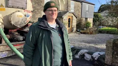 Karen Smith is standing near a water tanker and there is a pile of sandbags behind her in front of a row of houses. She is wearing an anorak and a woolly hat and a slight smile. 