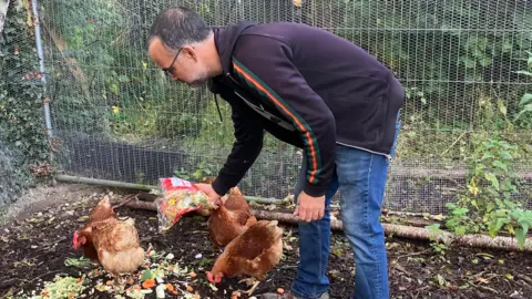 A man wearing jeans and a sports' top stoops over a group of 6 hens. He's emptying a bag of stir fry veg which has passed its "use by" date. The hens bend their heads in unison to pick at the scraps. In the background is fencing which forms part of a secure enclosure for the birds.
