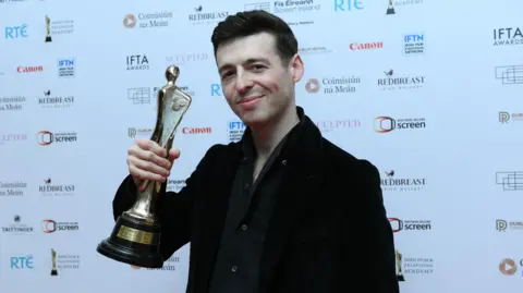 Charles McQuillan/Getty Images Anthony Boyle, a man with short, dark hair, holds up his Lead Actor award at the 2026 Irish Film & Television (IFTA) Awards at the Dublin Royal Convention Centre. He is wearing a dark, open-necked shirt under a black suit jacket. The award is a gold statuette with a Celtic design and black base with a small gold plaque.
