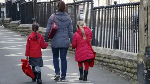 A generic picture showing the back of a woman walking along a street holding hands with two little girls, one with dark hair and the other with blonde hair. The girls are wearing school uniforms under red jackets. The woman is wearing a grey jacket and blue jeans. She has dark hair in a ponytail and is carrying a handbag. All have their backs to the camera. This is a stock picture