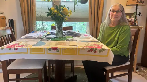 Sara Folger, 73, wears glasses and a green top, her long gray hair flowing down her back, as she sits at her kitchen table