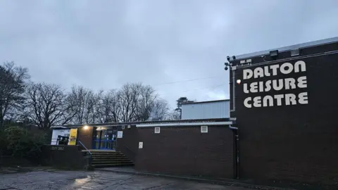 A brick building on a grey night with trees in the background. On the building is a large sign in white which says "Dalton Leisure Centre"