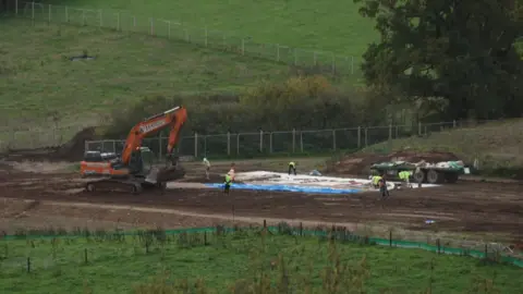A digger carrying out groundwork at Wolborough with staff in hi-vis working on a field
