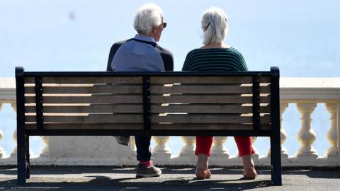 An elderly couple sit in the sunshine on the Hoe promenade overlooking the sea 