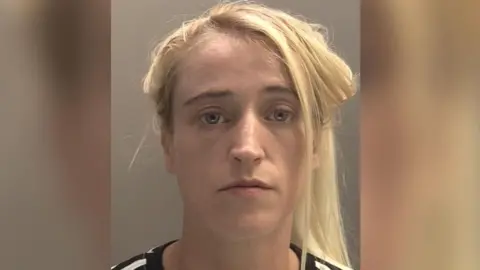 Police mugshot of Anya Foster looking impassively at the camera. She has long blonde hair which is loosely tied up. She has blue eyes and is wearing a black and white striped top.