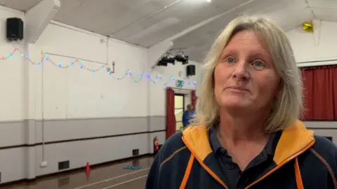 Allie is in the right hand side of the frame and looking out to the left hand side. She's wearing a black and orange hoodie with a black shirt underneath and has blonde medium-length hair. 