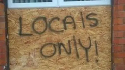 A wooden board has been placed over a PVC window frame. The words 'LOCALS ONLY!' is spray painted across it in black.  