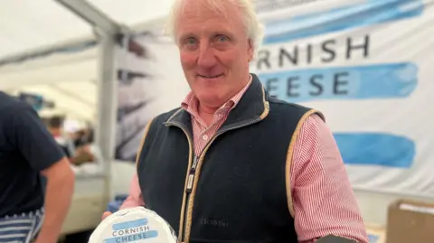 Phillip Stansfield, managing director of the Cornish Cheese Company, holds a wheel of cheese from his company inside a marquee at the Royal Cornwall Show. He is wearing a black gilet and a red and white stripped top.
