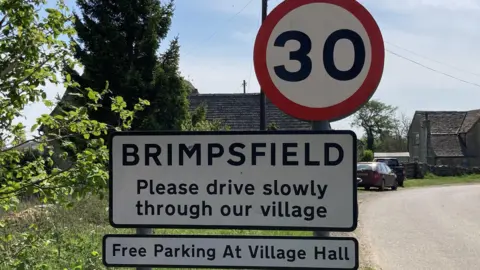 A photo showing a black and white welcome to Brimpsfield sign, with a 30 miles per hour sign above it. In the background is a road, car and a house