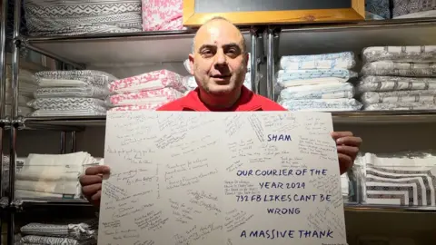 Sham, a delivery driver, holds up a large thank you card which is open and shows lots of messages and signatures from people thanking him for his work. In large letters a message reads: "our courier of the year 2024". Sham is wearing his black and red DPD drivers uniform and smiling