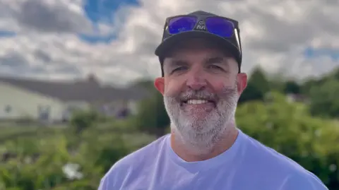 Damien Johnston standing outside with shrubs behind him. He is wearing a baseball cap, with dark glasses propped on its brim and a white T-shirt with Wembley 2 Wigmore written on it. He has a short cropped white beard and is smiling. 