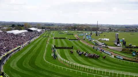 PA Media The riders - jockeys in colourful silks take a bend in front of a packed grandstand during Friday's racing at Aintree