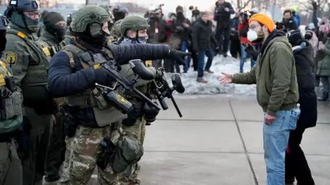 Reuters A federal agent wearing camouflage gestures towards a demonstrator wearing jeans and a green jumper and a face maskat a protest against the fatal shooting of Renee Nicole Good by a U.S. Immigration and Customs Enforcement (ICE) agent in Minneapolis, Minnesota