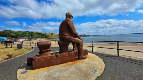 Figaro Large steel sculpture of a seated fisherman - a memorial to those lost at sea - on a the banks of a river stretching into the distance, framed by two piers as it meets the sea. To the left is a promenade with a low round pink building (an ice cream stand) backed by a bank of trees leading up to a large block of flats.