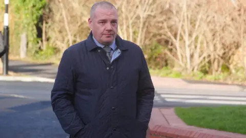 John Dance walking outside the court. He has a shaved head and is wearing a blue shirt and tie underneath a blue coat. He has his hands in his pockets and is looking into the camera.