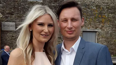 A couple pose for a picture - the woman on the left has long blonde hair and a light-pink dress. The man has short brown hair, a blue jacket and white shirt. There is an old stone building behind them.