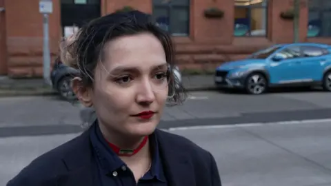 Maryam has brown hair with lighter colours tied up in the back. She's wearing red lipstick and has a navy shirt on. A street is behind her with parked cars. 