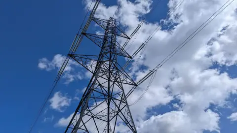 An overhead electricity pylon with cables attached against a blue sky with some clouds