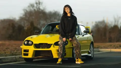 Kei car enthusiast Emma Bergeron sits on the bonnet of her mini sports car, a bright yellow modified Suzuki Cappuccino, in a car park next to a field.
