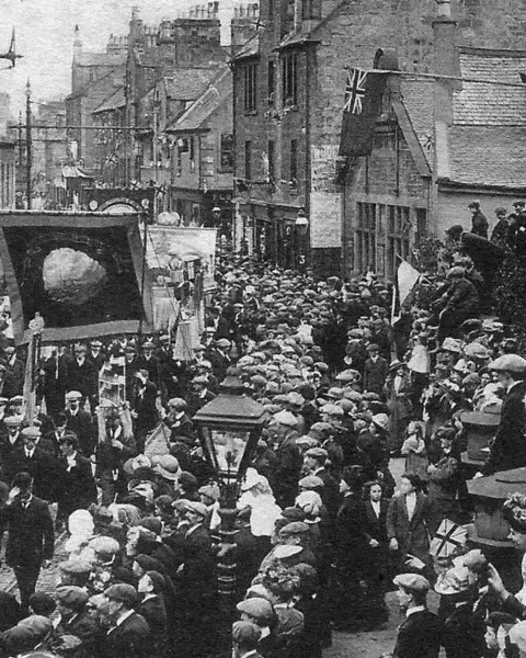 Kevin Green An old black and white photograph showing people, many in flat caps, watching a parade with banners. 