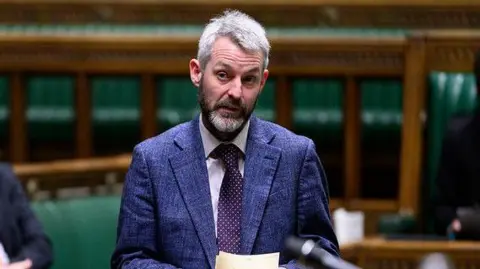 House of Commons A man, Will Forster MP, stood in a room with wooden walls and green cushioned seats. He is wearing a blue suit and speaking.