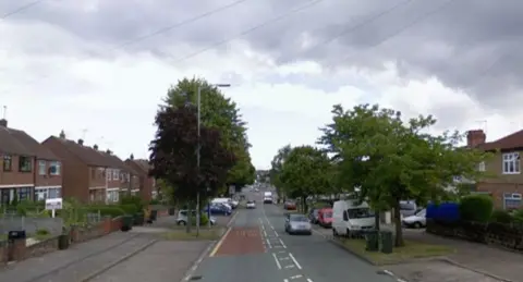 Google Clifford Bridge Road as shown on Google Street View. The road has houses on either side and some large trees and parked cars and a white van also parked. There are road markings down the centre of the street. 