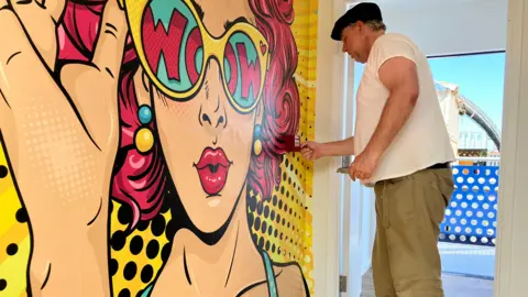 Jon Wright/BBC A man is standing in front of a wall which has a vibrant cartoon picture of a woman's face wearing sunglasses.