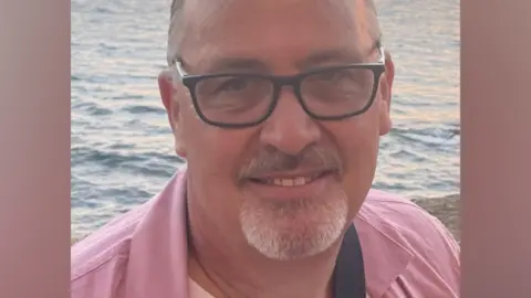 Matt Gaskin A man wearing glasses and a pink shirt is pictured outdoors by the sea, with water visible in the background.