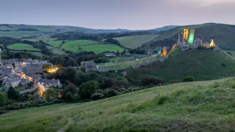 A long exposure shot of Corfe Castle lit up green and yellow with the village lights below