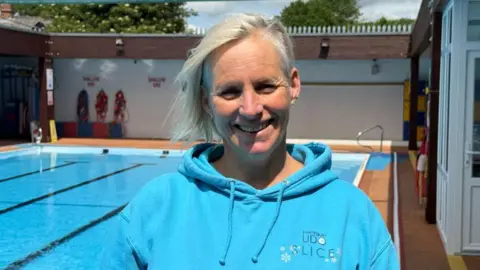Harriet has grey-blonde hair, styled to be longer on one side. She is smiling broadly for the camera, standing at the side of the pool with a blue fleece on.