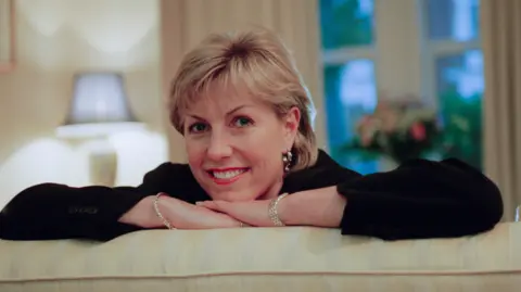 Jill Dando. She has short blonde hair and is wearing a black top and silver jewellery. She is leaning on the back of a sofa, with her chin resting on her hand. She is smiling at the camera. 