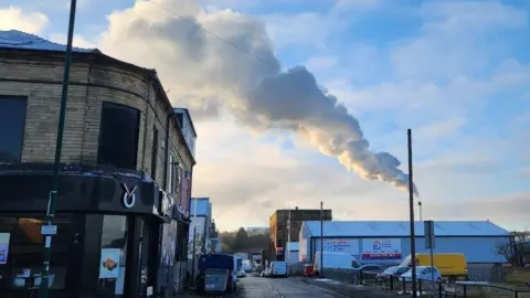 A street scene with various shops on one side and industrial buildings on the other. Dominating the skyline is a tall chimney with a huge plume of white smoke coming out of it and stretching across the sky. 
