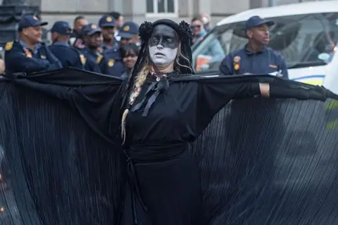 Getty Images Woman wearing all black and black and white face paint spreads open her cape as she protests with police behind her. 