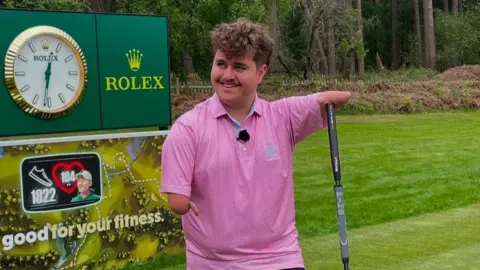 BBC Adam has under-developed arms. He is holding onto a golf club with his arm and wearing a pink golf shirt.