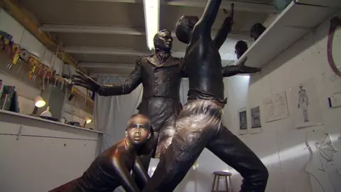 A bronze sculpture in a workshop. The walls are white and you can see tools hanging and drawings on them. The sculpture shows three figures – a shackled female slave kneeing down, an officer from the West Africa Squadron and a freed slave with his arms stretched to the sky.