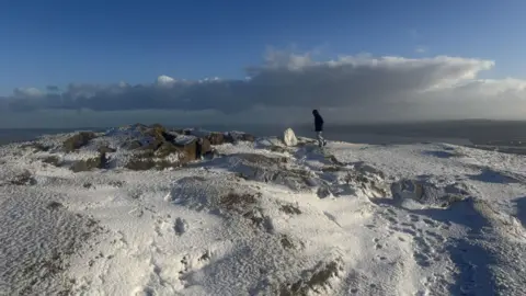 A man walks along the snowy summit of Cavehill. Footprints are visible in the snow