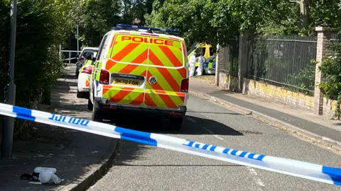 Two forensic investigators in white PPE can be seen on a residential street cordoned off by a blue and white police tape. A number of police vehicles are parked along the pavement.