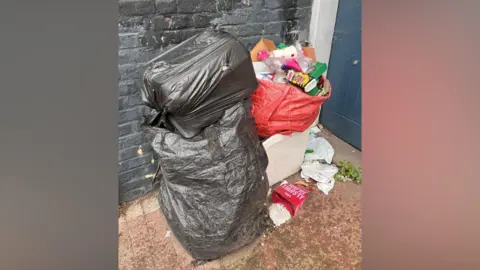 Phil Harrison/BBC A black rubbish bag stacked on top of another one. There is also a red bag filled with empty drinking bottles and cups. There are rubbish lying on the ground. 