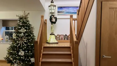 IWC Media Wooden staircase decorated for Christmas with a large lit tree with gold and white decorations, a large vintage-style lamp post with wreath, and miniature festive houses decorating the wall on the landing.