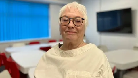 Sam Read/BBC Danielle Stone standing in a room, looking at the camera. She is wearing a cream top and has short fair hair with glasses and has earrings in both ears. The room behind her is slightly blurred and features a screen, a window and tables and chairs.