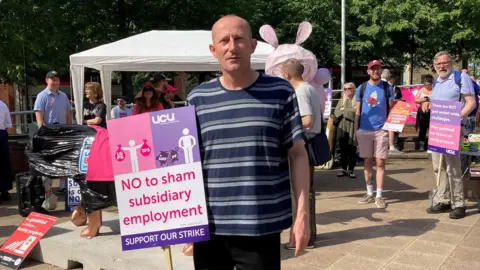 BBC/Naj Modak Ben Abel in a striped shirt stands in a paved area holding a purple and white UCU strike sign that reads "NO to sham subsidiary employment." In the background, people gather near a white canopy tent on a sunny day.