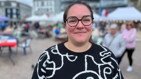 A woman wearing a black and white jumper. Her brown hair is tied back and she is wearing glasses. Behind her are people walking around and market stalls.
