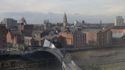 An aerial view of the black, futuristic Scale Lane bridge in Hull. A wooden walkway is visible along the bank of the river. The skyline of Hull city centre is visible in the background.